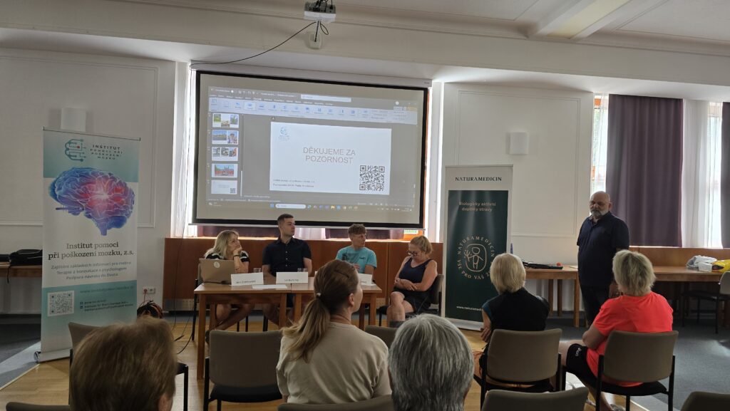 Dospělí na přednášce o podpoře poškozeného mozku s prezentací na velkém plátně, vedoucí diskusi odborníci v oblasti neurologie a rehabilitace.