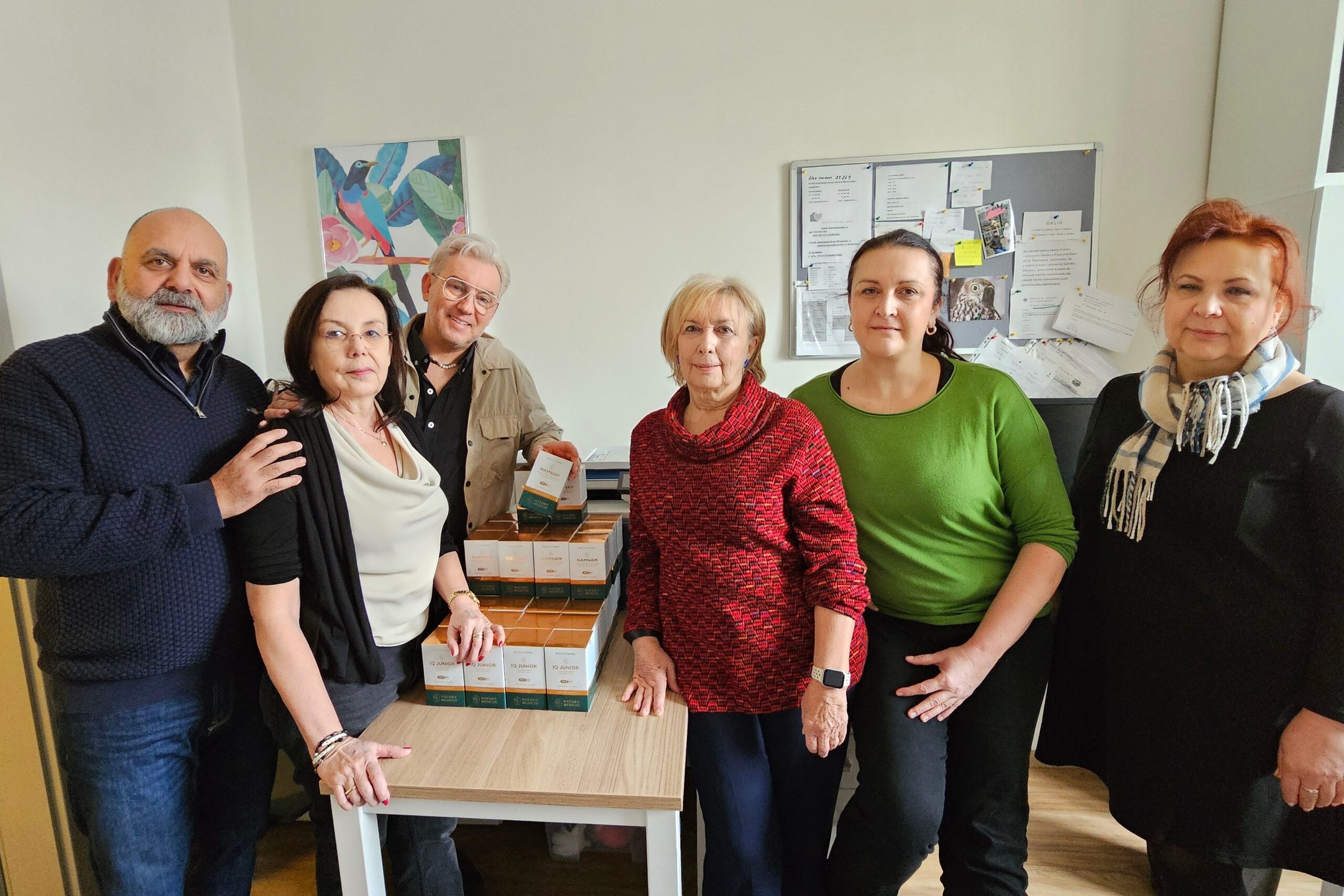 Popsaný obrázek: Skupina lidí stojí kolem stolu s produkty a dokumenty v kanceláři, zřejmě při charitativní nebo firemní akci, přičemž je zdůrazněna jejich spolupráce a společná činnost.