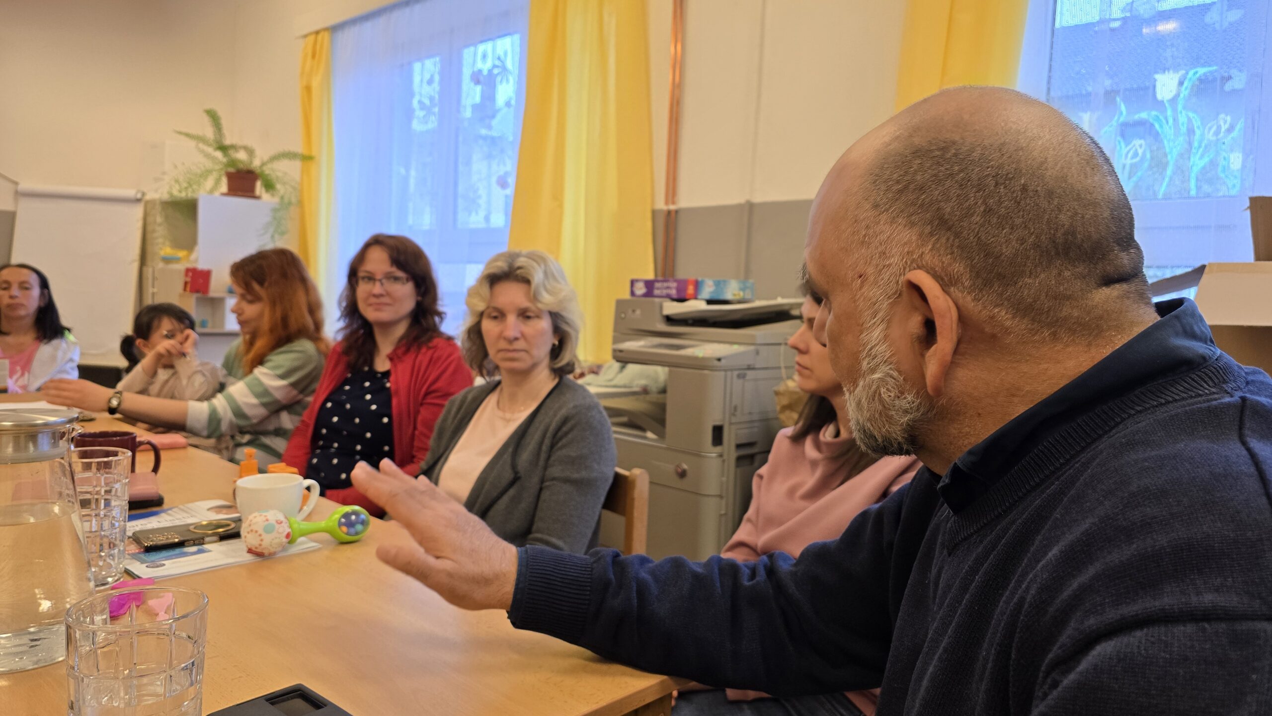 Kvalitní laboratorní technik Bohuslav Větrovský při práci na semináři o optických systémech. Skupina lidí sleduje prezentaci v učebně s pracovním prostředím a technikou, zaměřené na optické technologie a výzkum.